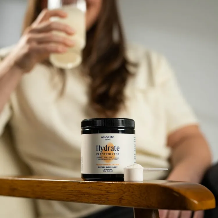 A woman drinking from a glass behind a jar of Ameo Life Hydrate Electrolytes and a scoop